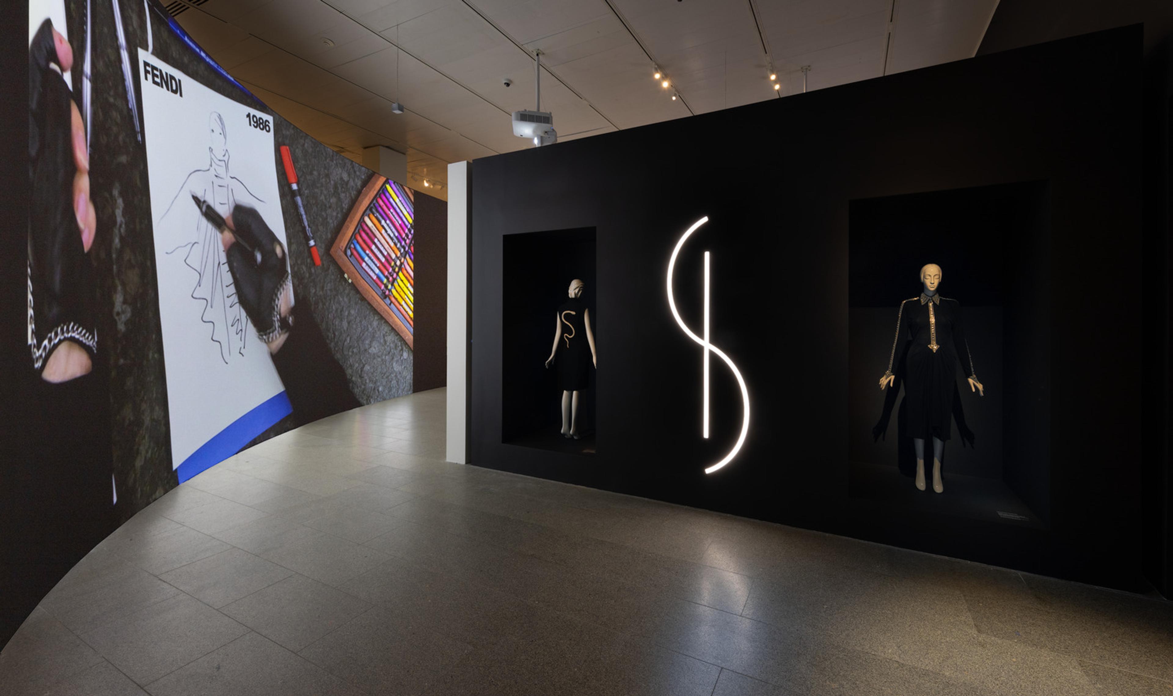 A sweeping view of a dark gallery featuring featuring two mannequins on the right and a projection of a gloved hand drawing a fashion sketch on the left.
